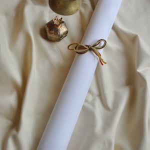 May include: A rolled white paper scroll tied with a brown and yellow twine bow. A small gold pomegranate-shaped container and a decorative brass lamp sit on a cream-colored fabric background. The scroll has wooden dowels.