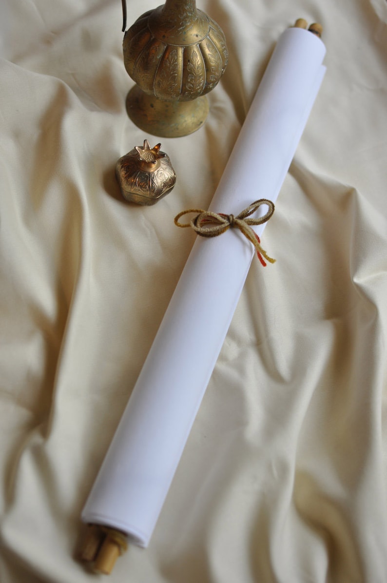 May include: A rolled white paper scroll tied with a brown and yellow twine bow. A small gold pomegranate-shaped container and a decorative brass lamp sit on a cream-colored fabric background. The scroll has wooden dowels.