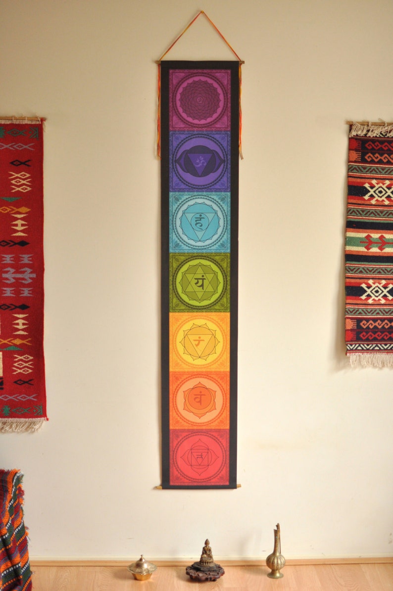 May include: A colorful wall tapestry depicting the seven chakras with their Sanskrit symbols and corresponding colors. The tapestry is hung on a white wall with two smaller rugs on either side.