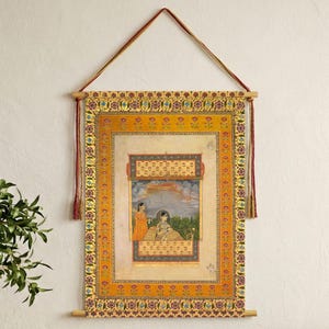 May include: An ornate, framed artwork featuring a traditional Indian miniature painting. The artwork is dominated by warm colors, including gold, orange, and red. The painting depicts two figures in a garden setting. The artwork is suspended by a wooden rod and a red cord.