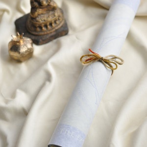 May include: A white scroll tied with a brown and red ribbon, resting on a white fabric surface. A small golden statue of a Buddha sits on a brown base to the left of the scroll.