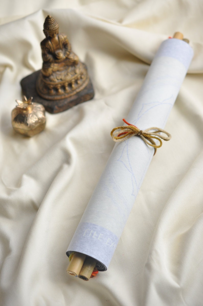 May include: A white scroll tied with a brown and red ribbon, resting on a white fabric surface. A small golden statue of a Buddha sits on a brown base to the left of the scroll.