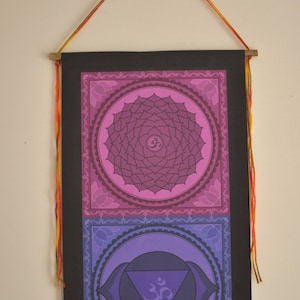 May include: A wall hanging with a black background and three colorful chakra symbols. The symbols are in shades of pink, purple, and blue. Each symbol is surrounded by a decorative border. The hanging has a string attached to the top for hanging.
