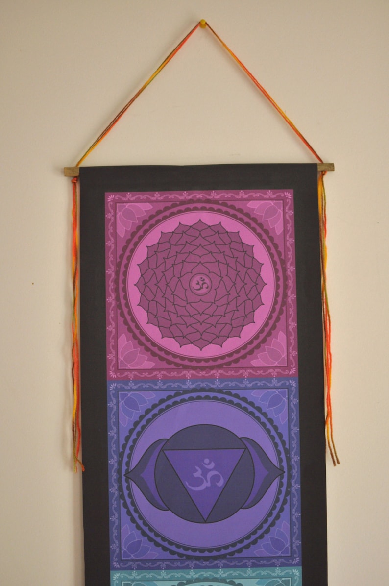 May include: A wall hanging with a black background and three colorful chakra symbols. The symbols are in shades of pink, purple, and blue. Each symbol is surrounded by a decorative border. The hanging has a string attached to the top for hanging.