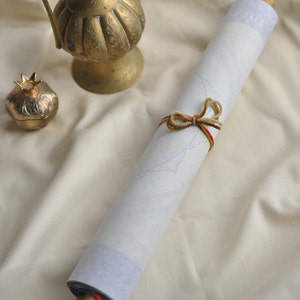 May include: A white scroll tied with brown and red string, rolled up and resting on a white fabric surface. A gold metal pitcher and a gold metal pomegranate ornament are also on the surface.