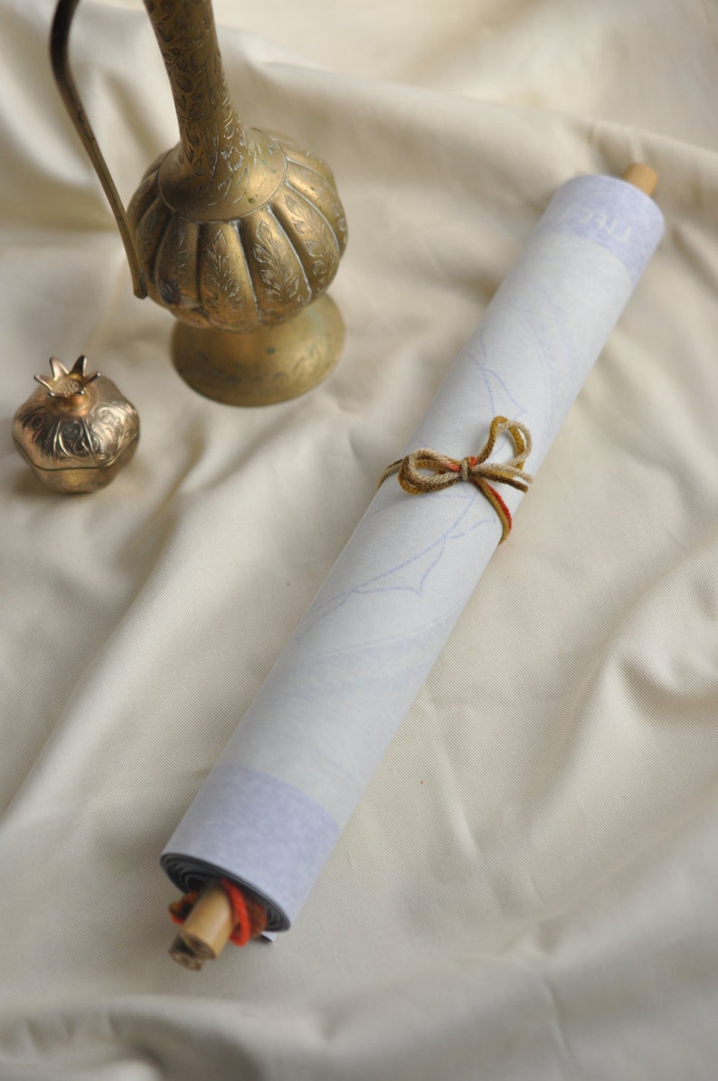 May include: A white scroll tied with brown and red string, rolled up and resting on a white fabric surface. A gold metal pitcher and a gold metal pomegranate ornament are also on the surface.