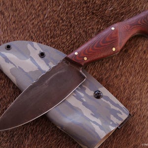 Puede incluir: Cuchillo artesanal con una hoja oscura y texturizada y un mango de madera. El mango es de un rico color marrón rojizo, acentuado con pasadores de latón. El cuchillo descansa sobre una funda con estampado de camuflaje.