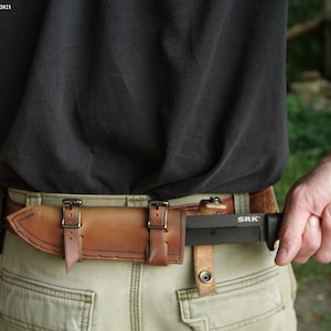 May include: A brown leather sheath with two buckles on each side is attached to a brown belt. A black knife with a black handle and the text "SRK" is in the sheath.