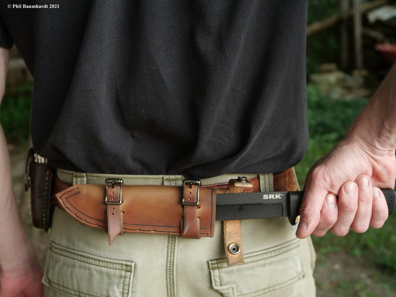 May include: A brown leather sheath with two buckles on each side is attached to a brown belt. A black knife with a black handle and the text "SRK" is in the sheath.