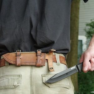 May include: A person wearing a black t-shirt and khaki pants with a brown leather belt and a knife sheath. The knife is black with a black handle and the text "SRK" on the blade.