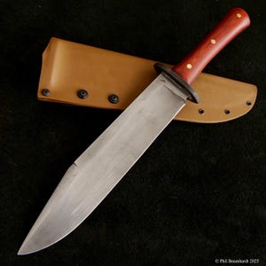 Könnte beinhalten: Ein großes, handgefertigtes Messer mit polierter Edelstahlklinge und dunklem Holzgriff. Der Griff ist mit Messingnieten befestigt. Das Messer liegt auf einer hellbraunen Lederscheide. Die Klinge ist etwa 30 cm lang.