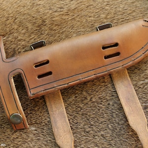 May include: A brown leather sheath with two straps and a metal buckle. The sheath has three cutouts on the front and is designed to hold a knife.