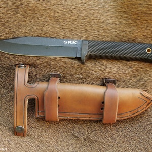 May include: A black knife with a brown leather sheath. The knife has a black handle with a textured grip and the text "SRK" on the blade. The sheath has a belt loop and two straps.