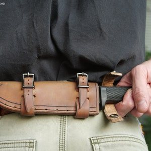 May include: A brown leather knife sheath with two straps and buckles. The sheath is attached to a belt and a knife is partially visible.
