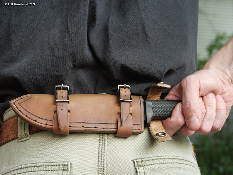 May include: A brown leather knife sheath with two straps and buckles. The sheath is attached to a belt and a knife is partially visible.