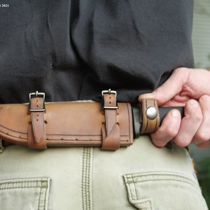 May include: A brown leather sheath with two straps and a metal buckle is attached to a belt. A black handled knife is secured in the sheath.