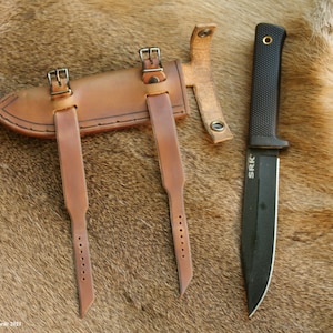 May include: A brown leather sheath with two straps and buckles, designed to hold a knife. A black knife with a black handle and the text "SRK" is shown next to the sheath.