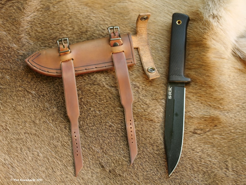 May include: A brown leather sheath with two straps and buckles, designed to hold a knife. A black knife with a black handle and the text "SRK" is shown next to the sheath.
