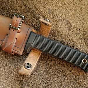 May include: A brown leather sheath with a buckle and a black knife with a textured handle. The knife is partially inserted into the sheath.