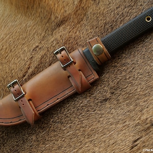 May include: A brown leather knife sheath with two straps and buckles. The sheath is designed to fit a knife with a black handle and a textured blade.