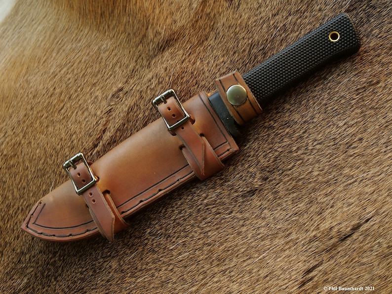 May include: A brown leather knife sheath with two straps and buckles. The sheath is designed to fit a knife with a black handle and a textured blade.