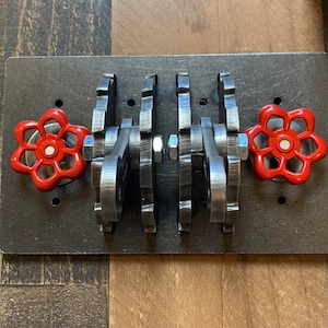 May include: A black metal wall plate with two red industrial-style valve handles. The handles are attached to a series of gears that are visible behind the handles.