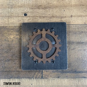 May include: A rusty brown metal gear with a black background. The gear is attached to a black square wooden plaque with four black screws.