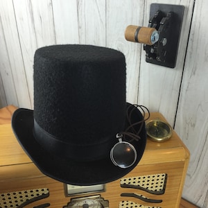 Op de afbeelding: Een zwarte hoge hoed met een bruin leren bandje en een vergrootglas hanger zit bovenop een vintage houten radio met een gouden wijzerplaat. Een houten schakelaar met tandwielen is gemonteerd op de muur achter de radio.