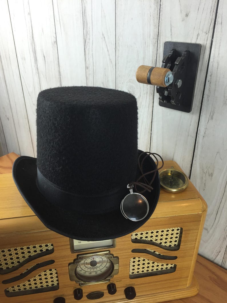 Op de afbeelding: Een zwarte hoge hoed met een bruin leren bandje en een vergrootglas hanger zit bovenop een vintage houten radio met een gouden wijzerplaat. Een houten schakelaar met tandwielen is gemonteerd op de muur achter de radio.
