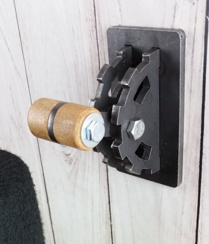 Op de afbeelding: Een zwarte metalen wandschakelaar met een houten handvat en een tandwielvormig ontwerp. De schakelaar is gemonteerd op een witte houten muur.