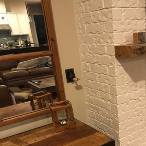 Op de afbeelding: Een witte bakstenen muur met een houten plank en een rustieke houten lantaarn. De lantaarn is gemaakt van hout en heeft een glazen paneel. De lantaarn staat op een houten tafel.