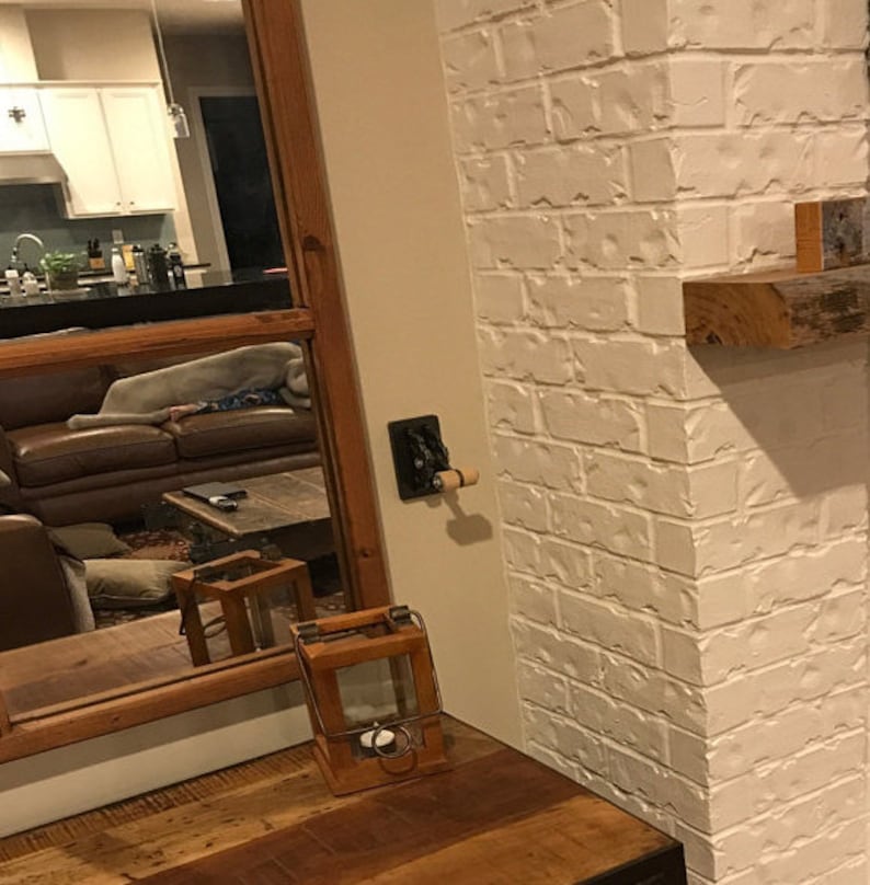 Op de afbeelding: Een witte bakstenen muur met een houten plank en een rustieke houten lantaarn. De lantaarn is gemaakt van hout en heeft een glazen paneel. De lantaarn staat op een houten tafel.