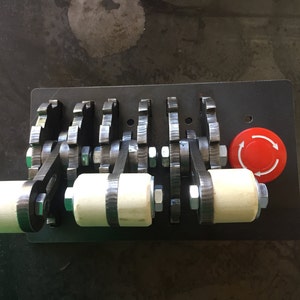 May include: A metal panel with four black levers with white handles and a red button with a white circular arrow symbol. The levers are connected by gears.