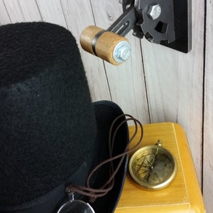 Op de afbeelding: Een zwarte hoge hoed met een bruin koord en een vergrootglas eraan bevestigd. De hoed ligt op een houten radio met een kompas en een steampunk-achtige wandschakelaar met een houten handvat.