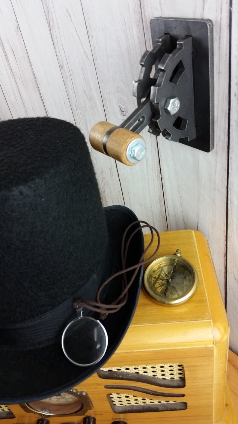 Op de afbeelding: Een zwarte hoge hoed met een bruin koord en een vergrootglas eraan bevestigd. De hoed ligt op een houten radio met een kompas en een steampunk-achtige wandschakelaar met een houten handvat.