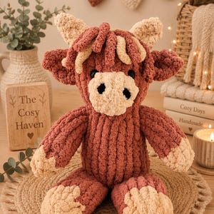 May include: A handmade, plush Highland cow stuffed animal in shades of brown and cream. The cow has a textured, chunky knit appearance, with a black nose and eyes. The text "The Cosy Haven" is on a wooden sign.