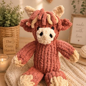 May include: A handmade, plush Highland cow stuffed animal in shades of dusty rose and cream. The cow has a textured, ribbed body, floppy ears, and a fluffy fringe. The Cozy Haven sign is visible in the background.