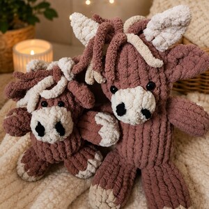 May include: Two handmade, plush cow stuffed animals. The cows are a dusty rose color with cream-colored accents on the ears, muzzle, and hooves. They have black button eyes and are sitting on a cream-colored, textured blanket.