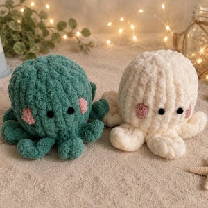 Op de afbeelding: Twee gehaakte octopus speelgoed, een in teal en een in cr&egrave;me, met zwarte ogen en roze wangen, liggen op een zanderig oppervlak. De teal octopus heeft een donkerdere groene tint, terwijl de cr&egrave;me octopus een zacht off-white is. Een vuurtoren en schelpen zijn ook zichtbaar.