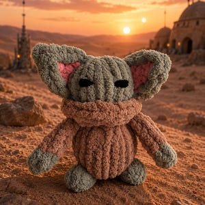 May include: A crocheted plush toy of a character with large ears, black eyes, and a tan-colored body. The toy is sitting on a sandy surface with a sunset in the background. The ears have pink inner linings.