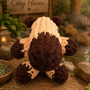 May include: A plush capybara toy crafted from chunky yarn, featuring a light beige body with dark brown accents on the ears, snout, and feet. The stuffed animal has round black eyes and a soft, textured appearance with a rounded, bulky shape.