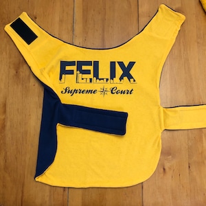 May include: Yellow dog vest with blue trim and a black patch on the shoulder. The vest has the text "FELIX Supreme Court" printed on the front.