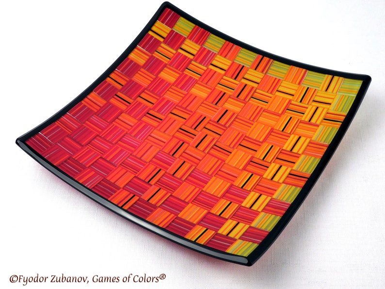 May include: A square glass serving dish with a black rim. The dish is decorated with a woven pattern of red, orange, and yellow glass strips.