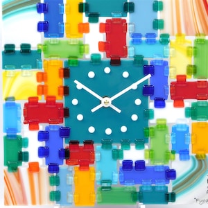 May include: A square wall clock with a teal center and colorful glass blocks in red, blue, green, and yellow. The clock has white hands and a white face with dots. The clock is a decorative item.