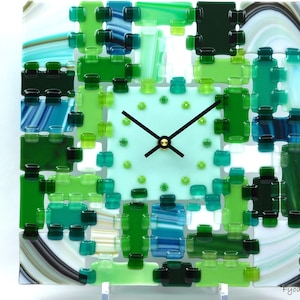 May include: Square glass clock with a light green face and black hands. The clock face is surrounded by a mosaic of green and blue glass pieces. The clock is supported by a clear stand. The clock is a decorative home decor item.