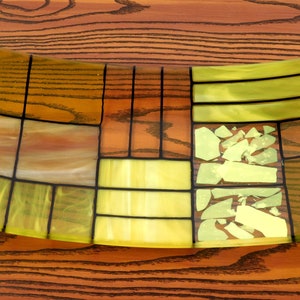 Puede incluir: Un plato de servir de vidrio curvo con un patr&oacute;n geom&eacute;trico de piezas de vidrio amarillo, verde y marr&oacute;n. El plato tiene un dise&ntilde;o moderno y abstracto.