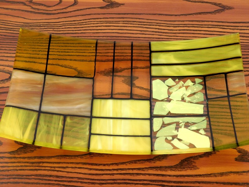 Puede incluir: Un plato de servir de vidrio curvo con un patr&oacute;n geom&eacute;trico de piezas de vidrio amarillo, verde y marr&oacute;n. El plato tiene un dise&ntilde;o moderno y abstracto.