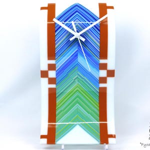 May include: A rectangular glass clock with a white face and white clock hands. The clock face features a chevron pattern in shades of blue and green. The clock is framed with white and brown accents. The clock is approximately 10 inches tall.