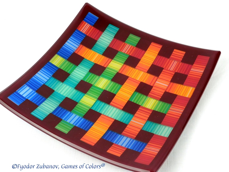 May include: A square glass plate with a woven pattern in shades of blue, green, yellow, orange, and red. The plate has a dark red border.