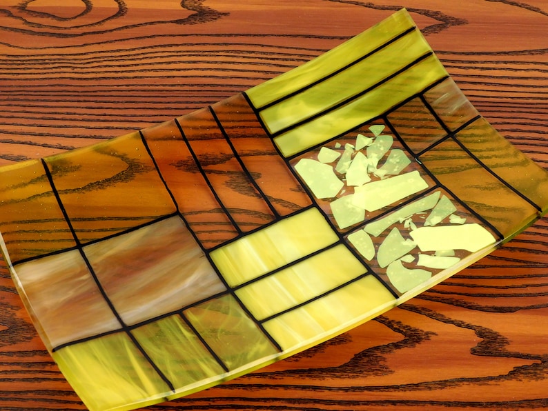Puede incluir: Un plato rectangular de vidrio con un patr&oacute;n geom&eacute;trico de piezas de vidrio amarillo y marr&oacute;n. El plato tiene un borde ligeramente curvado y est&aacute; hecho de vidrio emplomado.